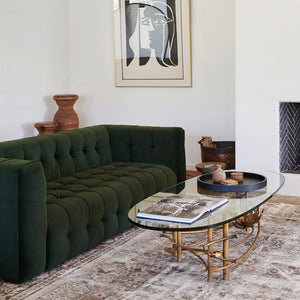 Green tufted velvet sofa in white room with vintage glass and guilded coffee table and abstract art on wall.