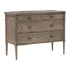 This dresser has a rectangular shape with three wide drawers and a weathered, rustic wood texture. The edges are softly rounded, and the small oval knobs have a smooth finish. Its slim, tapered legs feature subtle carved detailing, adding a refined touch. The overall look is simple yet elegant, with a pale, vintage-style finish.