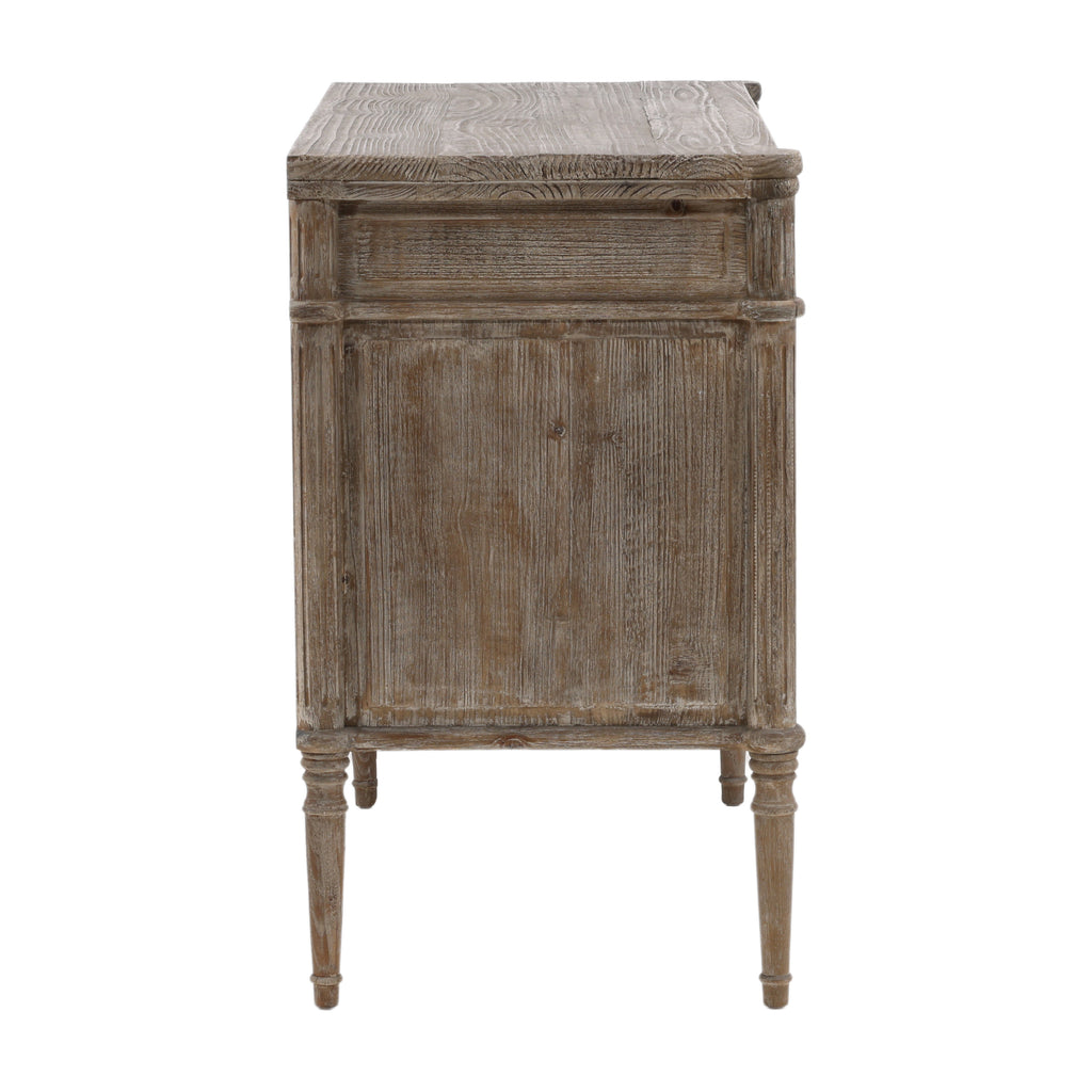 This dresser has a rectangular shape with three wide drawers and a weathered, rustic wood texture. The edges are softly rounded, and the small oval knobs have a smooth finish. Its slim, tapered legs feature subtle carved detailing, adding a refined touch. The overall look is simple yet elegant, with a pale, vintage-style finish.