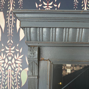 This mirror has a rectangular shape and is surrounded by a wooden frame that feels solid and smooth to the touch. The frame is painted in a soft, muted blue color and features subtle carved detailing along the top edge, giving it a slightly ornate appearance. The wood has a distressed finish, with small imperfections that add to its vintage charm.