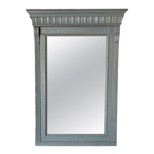 This mirror has a rectangular shape and is surrounded by a wooden frame that feels solid and smooth to the touch. The frame is painted in a soft, muted blue color and features subtle carved detailing along the top edge, giving it a slightly ornate appearance. The wood has a distressed finish, with small imperfections that add to its vintage charm.
