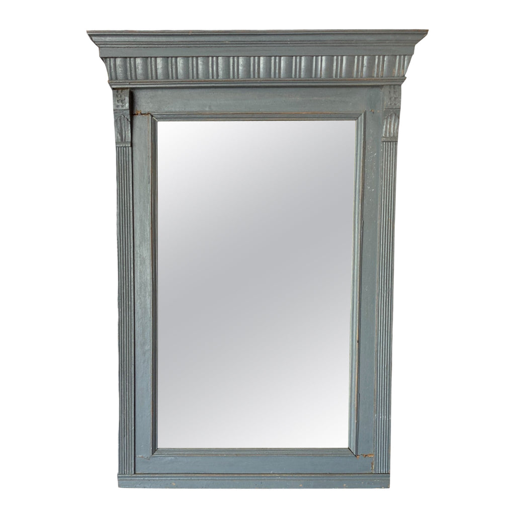 This mirror has a rectangular shape and is surrounded by a wooden frame that feels solid and smooth to the touch. The frame is painted in a soft, muted blue color and features subtle carved detailing along the top edge, giving it a slightly ornate appearance. The wood has a distressed finish, with small imperfections that add to its vintage charm.