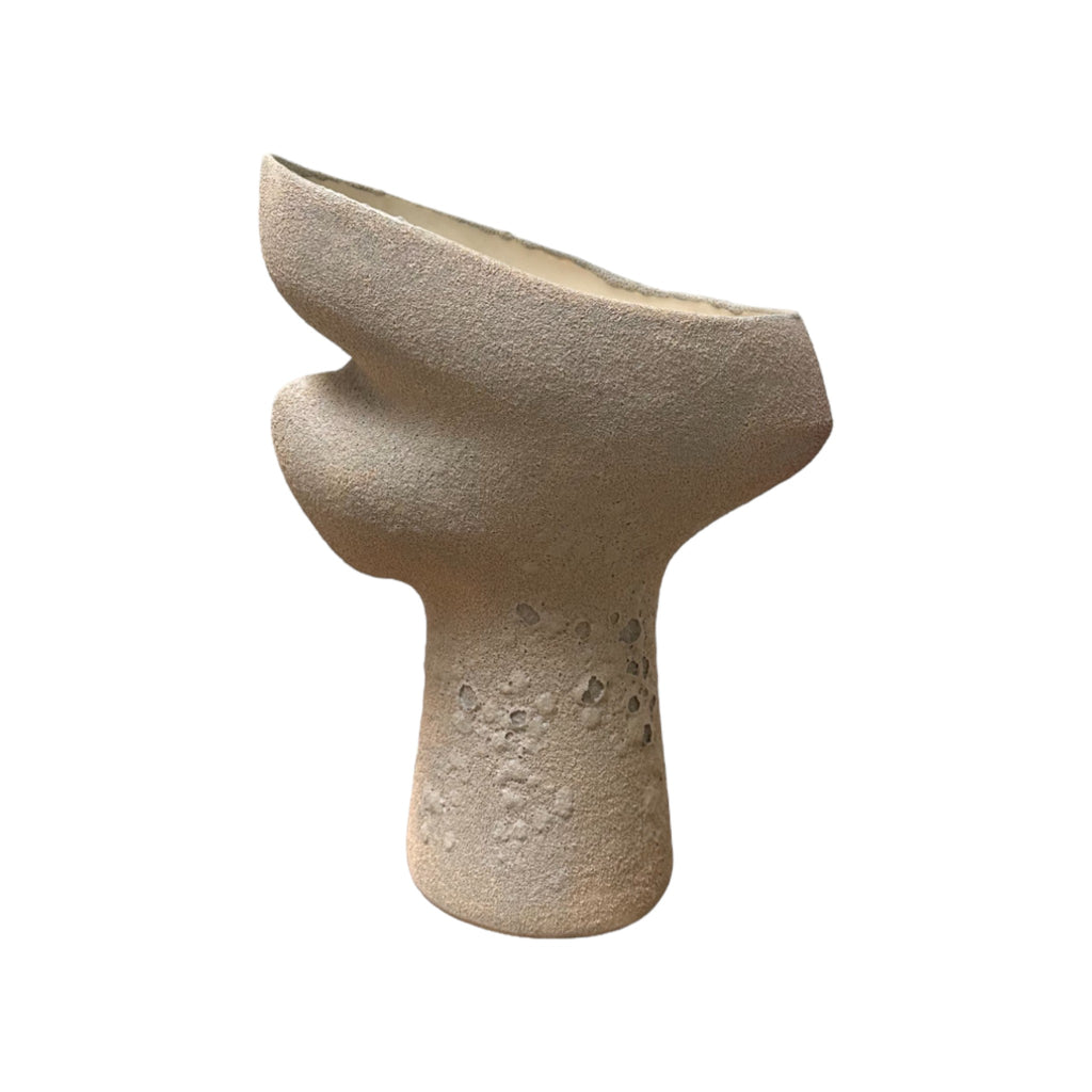 This sculpture is a handmade ceramic piece with a rough, sandy texture, resembling the feel of a coarse, natural stone. It has an abstract shape with a broad, curved top that narrows into a more cylindrical base. The surface features small indentations and variations, giving it a weathered, organic feel. The overall form is asymmetrical, with flowing, uneven curves that suggest movement and modern design.