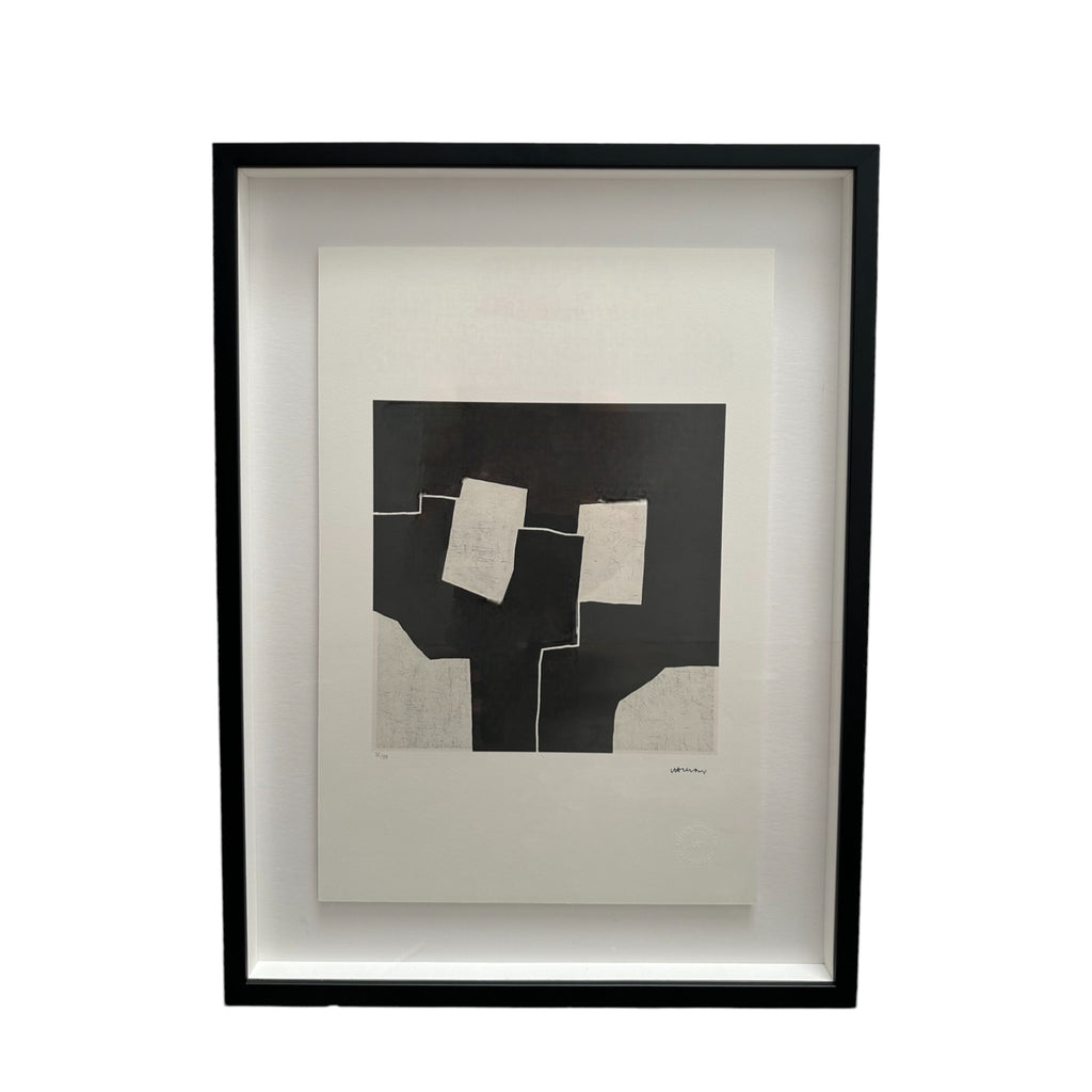 This piece is an abstract artwork by Eduardo Chillida, consisting of large black and white geometric shapes. The central area of the print features several irregular white rectangles arranged on a solid black background, giving the impression of overlapping forms. The design is minimalist, with sharp edges and contrasting tones that create a bold visual effect. It’s framed in a sleek black frame, adding a modern touch to the presentation.