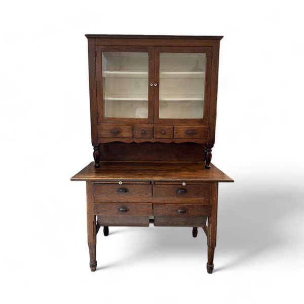 This vintage kitchen hutch is a tall, two-part wooden piece of furniture with a rich, dark finish. The upper section consists of a glass-fronted cabinet with two doors that open to reveal shelves for storing or displaying items. Below the glass cabinet, there are three small drawers, each with a metal pull, and a decorative scalloped trim. The lower section features a wide surface for working or serving, with four additional drawers beneath it, supported by four slender, turned legs. The overall design is r