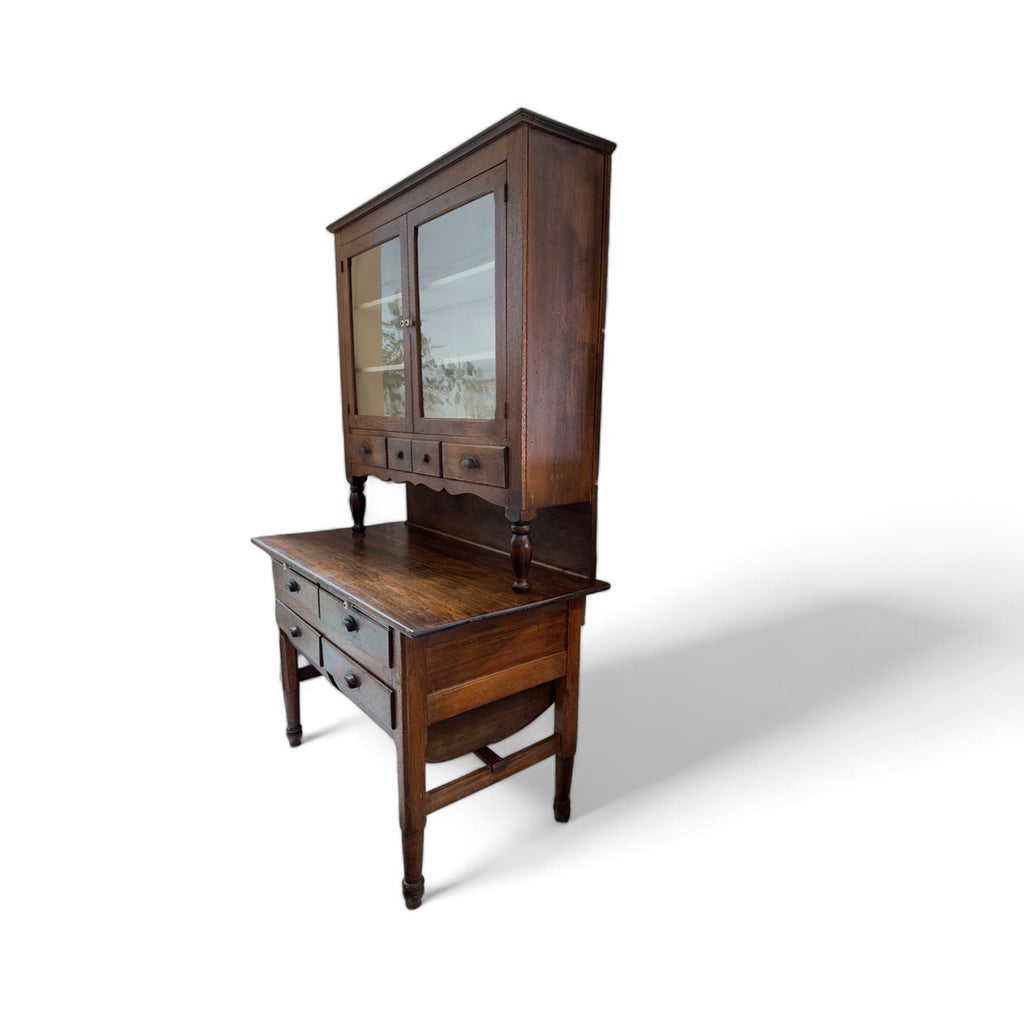 This vintage kitchen hutch is a tall, two-part wooden piece of furniture with a rich, dark finish. The upper section consists of a glass-fronted cabinet with two doors that open to reveal shelves for storing or displaying items. Below the glass cabinet, there are three small drawers, each with a metal pull, and a decorative scalloped trim. The lower section features a wide surface for working or serving, with four additional drawers beneath it, supported by four slender, turned legs. The overall design is r