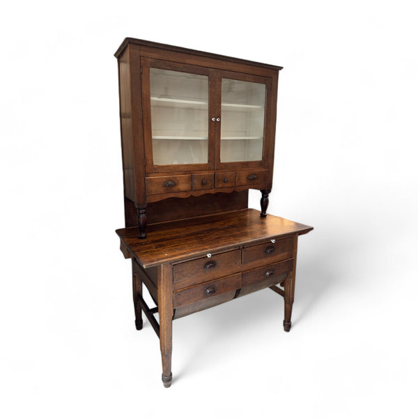 This vintage kitchen hutch is a tall, two-part wooden piece of furniture with a rich, dark finish. The upper section consists of a glass-fronted cabinet with two doors that open to reveal shelves for storing or displaying items. Below the glass cabinet, there are three small drawers, each with a metal pull, and a decorative scalloped trim. The lower section features a wide surface for working or serving, with four additional drawers beneath it, supported by four slender, turned legs. The overall design is r