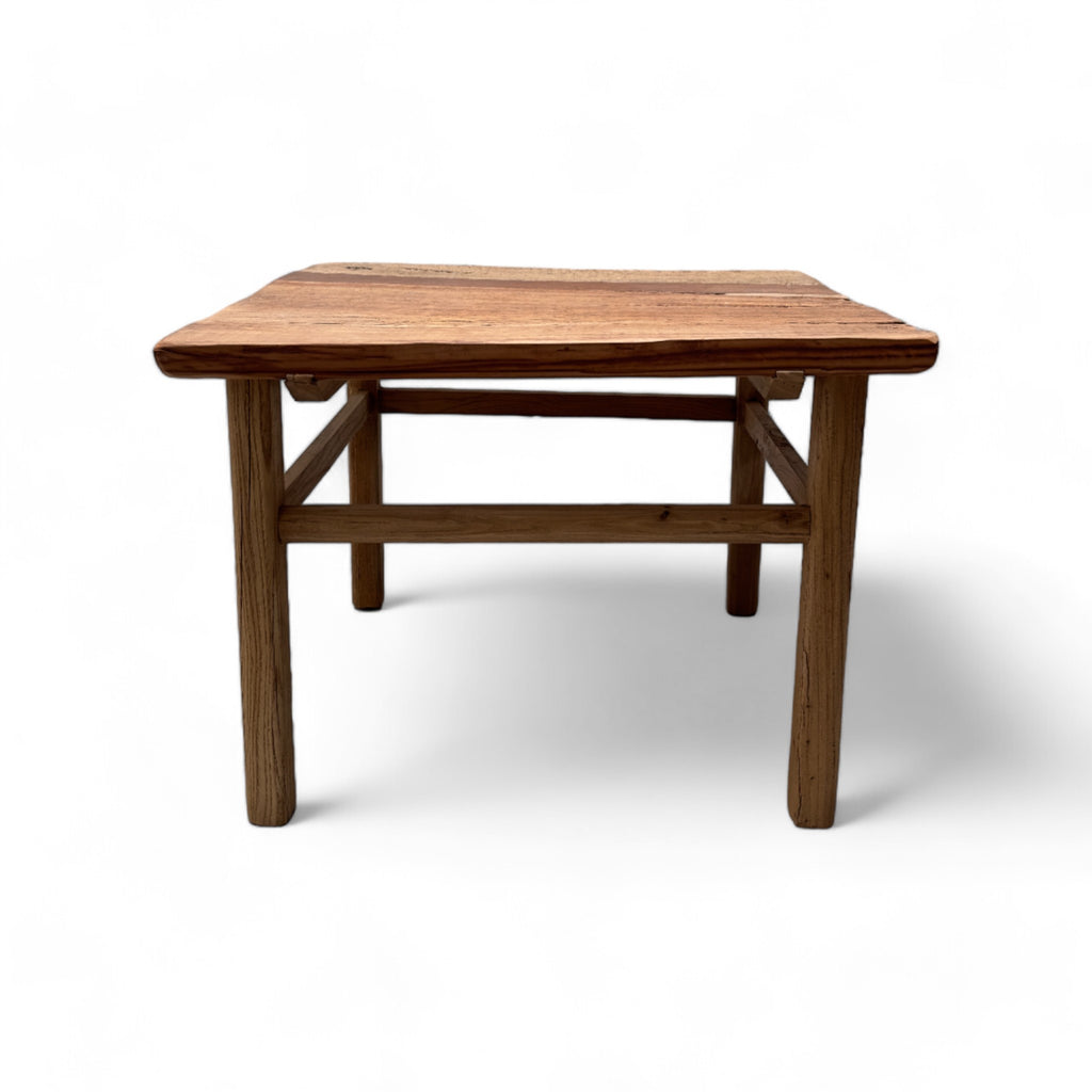 This coffee table is a square, low-profile piece made entirely of solid elm wood, giving it a natural, rustic appearance. The tabletop has a slightly uneven texture, with visible wood grain and color variations that add to its organic charm. The legs are thick and cylindrical, providing sturdy support, and are connected by simple wooden crossbars near the bottom. The overall design is straightforward and sturdy, with a warm, weathered finish that gives it a cozy, rustic feel.