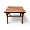 This coffee table is a square, low-profile piece made entirely of solid elm wood, giving it a natural, rustic appearance. The tabletop has a slightly uneven texture, with visible wood grain and color variations that add to its organic charm. The legs are thick and cylindrical, providing sturdy support, and are connected by simple wooden crossbars near the bottom. The overall design is straightforward and sturdy, with a warm, weathered finish that gives it a cozy, rustic feel.