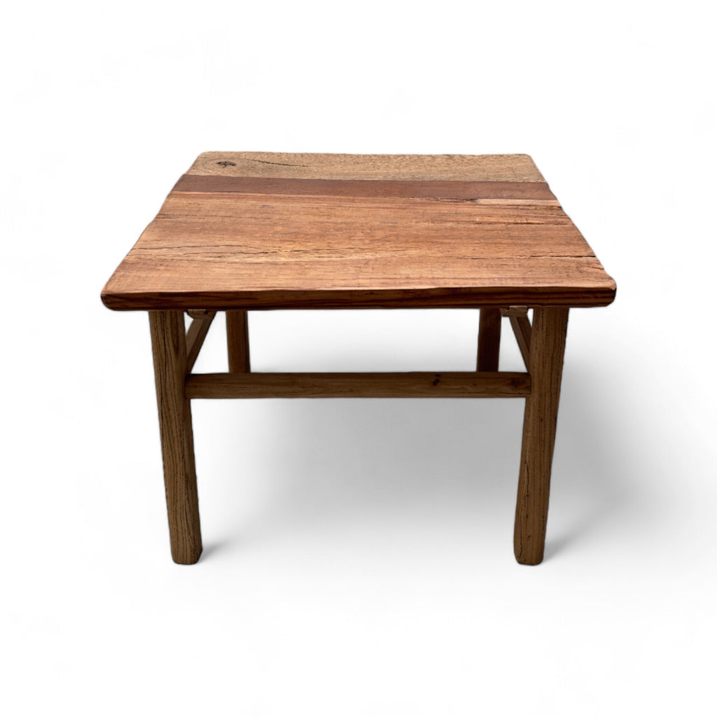 This coffee table is a square, low-profile piece made entirely of solid elm wood, giving it a natural, rustic appearance. The tabletop has a slightly uneven texture, with visible wood grain and color variations that add to its organic charm. The legs are thick and cylindrical, providing sturdy support, and are connected by simple wooden crossbars near the bottom. The overall design is straightforward and sturdy, with a warm, weathered finish that gives it a cozy, rustic feel.