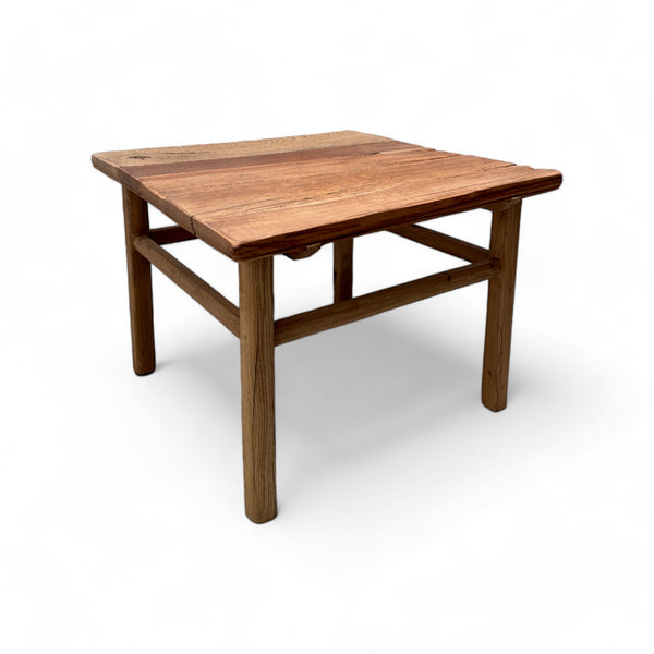 This coffee table is a square, low-profile piece made entirely of solid elm wood, giving it a natural, rustic appearance. The tabletop has a slightly uneven texture, with visible wood grain and color variations that add to its organic charm. The legs are thick and cylindrical, providing sturdy support, and are connected by simple wooden crossbars near the bottom. The overall design is straightforward and sturdy, with a warm, weathered finish that gives it a cozy, rustic feel.