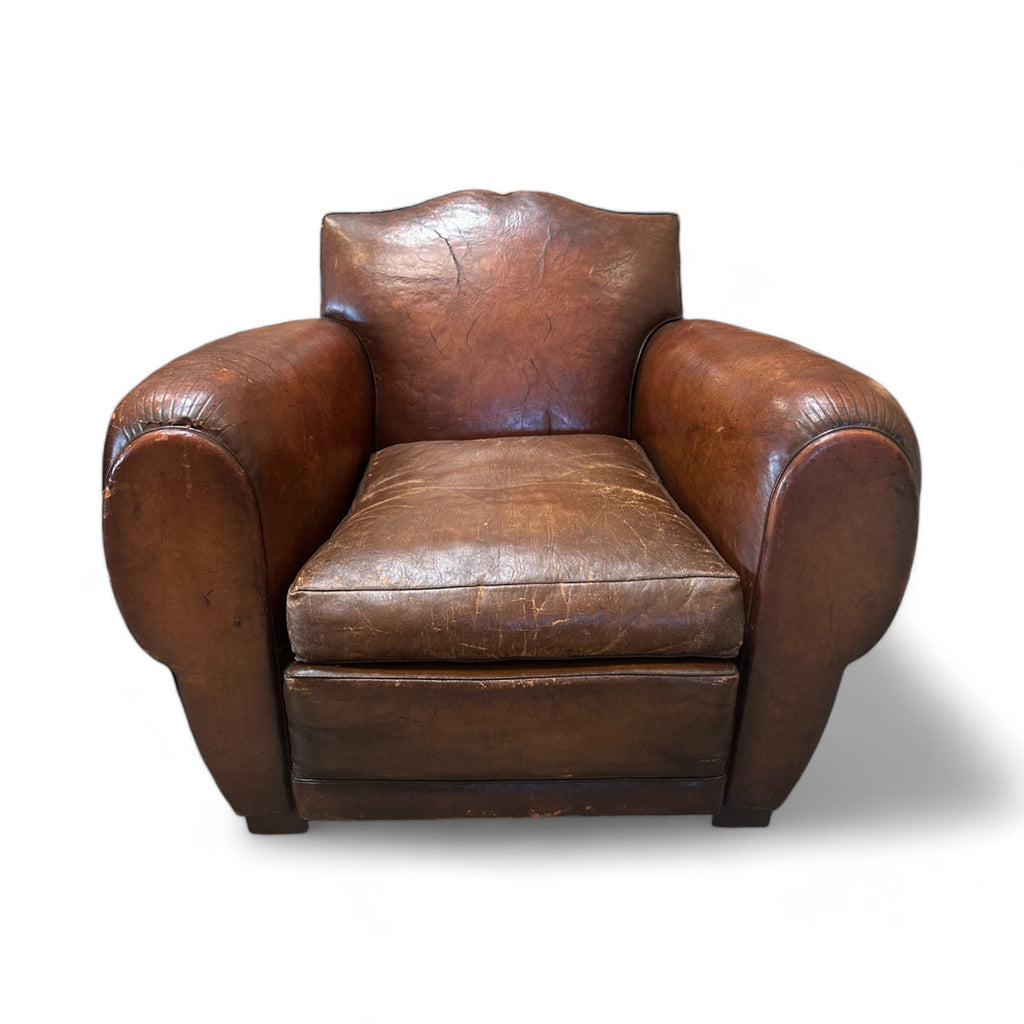 These chairs are sturdy, with a soft, worn leather surface that feels smooth in some areas but textured and creased in others, showing their age and use. The backrest is shaped like a subtle mustache, curving gently at the top, while the arms are wide and round, like soft, cushioned barrels that extend outward. Sitting in them, you’d notice they are large and enveloping, offering a sense of comfort and security, as the seat is deep and spacious, with firm padding beneath the aged leather.