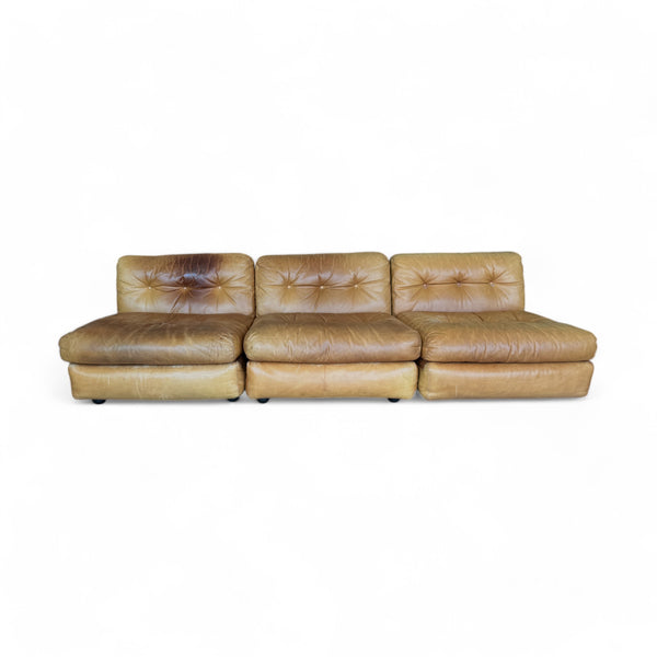 The set has a soft, aged leather in a warm tan shade that shows natural wear, giving it character. The seating is plump and low to the ground, with deep, buttoned tufting on the backrests and seats, creating a cozy, cushioned appearance. The sofa is modular, broken into three sections that align in a straight row, while the armchair has a similar, inviting design with rounded edges. The overall look feels both vintage and comfortably worn, like a well-loved, inviting piece of furniture.