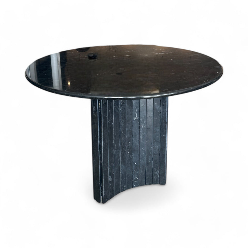 The table has a sturdy, elegant feel, with a cool, polished surface that’s smooth except for faint natural veining in the stone. Its base is uniquely shaped like a triangle, but instead of flat sides, each side curves gently inward, giving it a soft, sculpted form. Lining this triangular base are slim, vertical columns that add texture and depth, creating a sophisticated balance between solidity and subtle detailing.