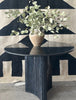 The table has a sturdy, elegant feel, with a cool, polished surface that’s smooth except for faint natural veining in the stone. Its base is uniquely shaped like a triangle, but instead of flat sides, each side curves gently inward, giving it a soft, sculpted form. Lining this triangular base are slim, vertical columns that add texture and depth, creating a sophisticated balance between solidity and subtle detailing.