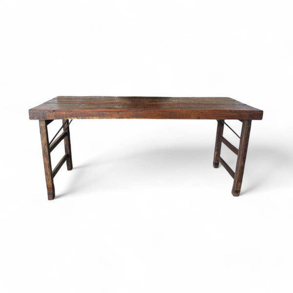 This table has a rectangular shape with a smooth, weathered wooden surface that feels sturdy and slightly textured under your fingers, showing the grain and natural imperfections of aged wood. The legs are simple and straight, connected by horizontal bars on each side, giving it a balanced, practical design. It’s solid to the touch, yet the clean lines make it feel light and adaptable to different spaces.