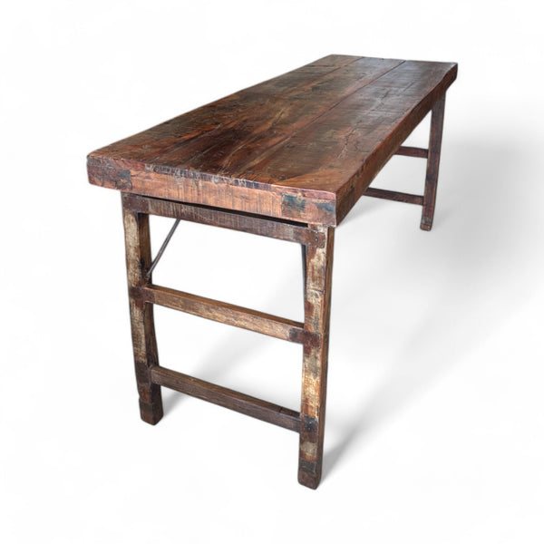 This table has a rectangular shape with a smooth, weathered wooden surface that feels sturdy and slightly textured under your fingers, showing the grain and natural imperfections of aged wood. The legs are simple and straight, connected by horizontal bars on each side, giving it a balanced, practical design. It’s solid to the touch, yet the clean lines make it feel light and adaptable to different spaces.