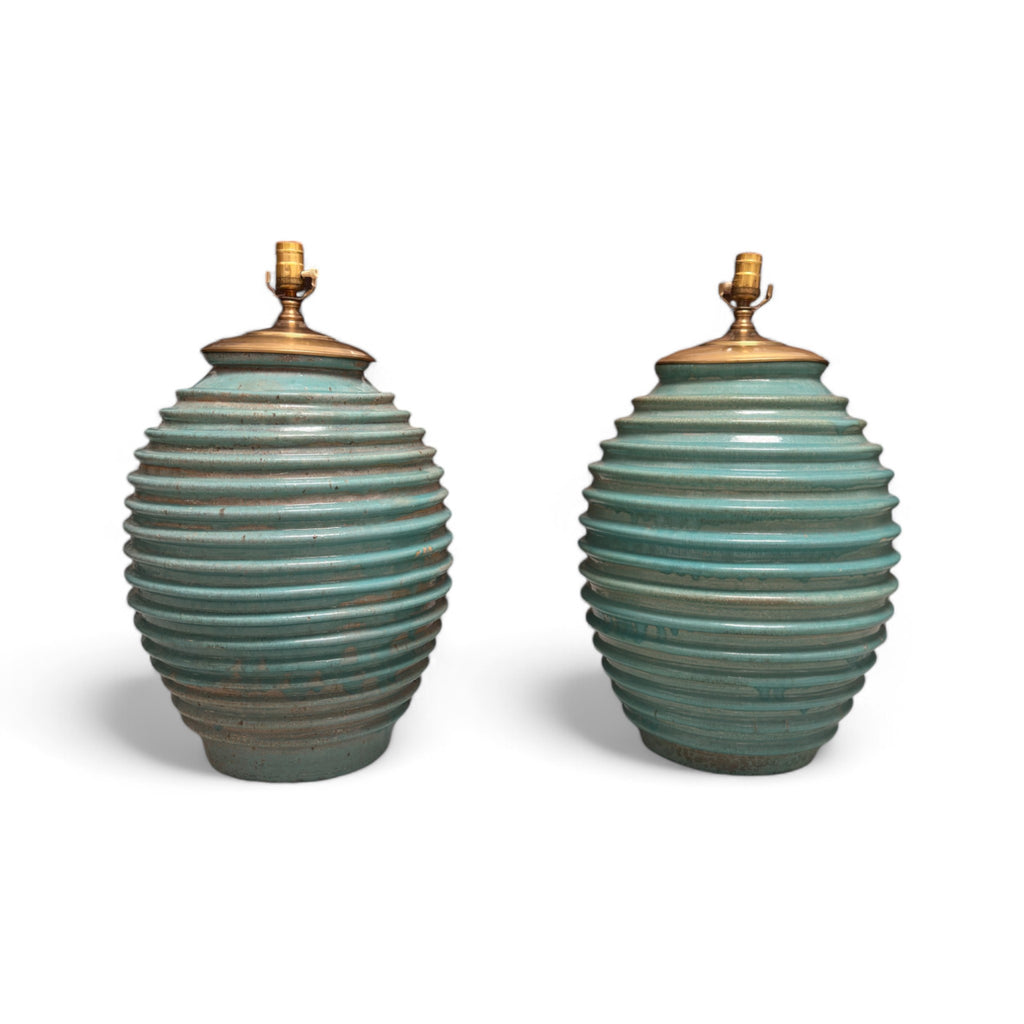 These lamps are shaped like large, rounded ovals with a ribbed texture running horizontally around the entire body. They have a soft green color with brass hardware at the top where the lightbulb fits in. The lamps feel slightly textured due to the ribbing, and the base is wide and sturdy.