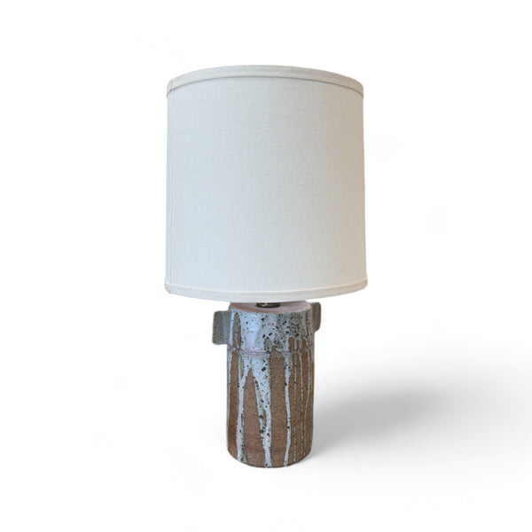 This lamp has a tall, cylindrical base with a smooth texture, but some areas feel as though they have vertical streaks where a glaze dripped down, leaving a rougher, uneven surface. There are small, flat, rectangular wings on either side of the base. The shade is tall and cylindrical, covered in a soft fabric, creating a smooth and clean finish.