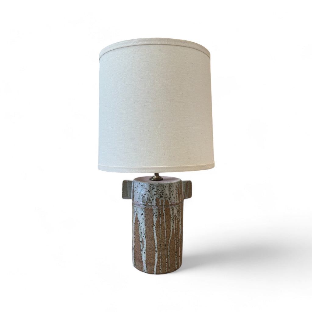 This lamp has a tall, cylindrical base with a smooth texture, but some areas feel as though they have vertical streaks where a glaze dripped down, leaving a rougher, uneven surface. There are small, flat, rectangular wings on either side of the base. The shade is tall and cylindrical, covered in a soft fabric, creating a smooth and clean finish.