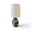 This lamp has a round, slightly bulging base that feels like textured clay, with some smooth raised lines, almost like the surface of a beehive. There are areas with circular, slightly rougher patches that feel like black paint. The lampshade is cylindrical, tall, and smooth, covered in a soft fabric, giving it a clean, minimalist feel.