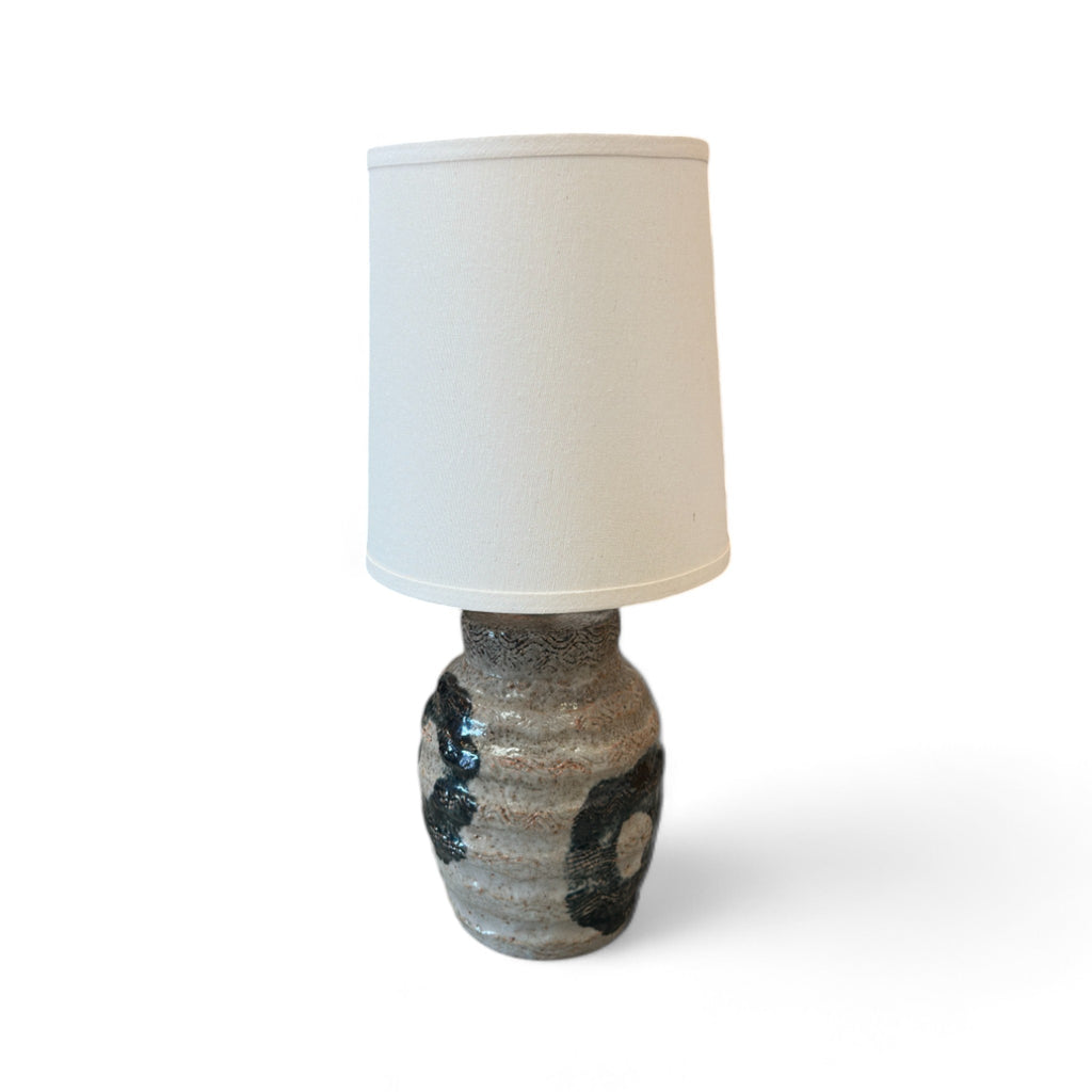 This lamp has a round, slightly bulging base that feels like textured clay, with some smooth raised lines, almost like the surface of a beehive. There are areas with circular, slightly rougher patches that feel like black paint. The lampshade is cylindrical, tall, and smooth, covered in a soft fabric, giving it a clean, minimalist feel.