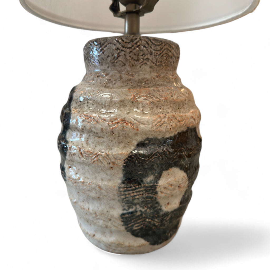 This lamp has a round, slightly bulging base that feels like textured clay, with some smooth raised lines, almost like the surface of a beehive. There are areas with circular, slightly rougher patches that feel like black paint. The lampshade is cylindrical, tall, and smooth, covered in a soft fabric, giving it a clean, minimalist feel.