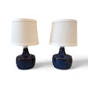 These are two small, matching lamps with round bases that feel smooth and glossy, likely ceramic. They have raised circular patterns on the sides that feel like little wheels or sunbursts. The lampshades are simple, small, and conical, covered in soft fabric. While the lamps themselves are identical in shape and texture, their plug-in cords are different, so they don’t completely match.