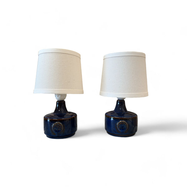 These are two small, matching lamps with round bases that feel smooth and glossy, likely ceramic. They have raised circular patterns on the sides that feel like little wheels or sunbursts. The lampshades are simple, small, and conical, covered in soft fabric. While the lamps themselves are identical in shape and texture, their plug-in cords are different, so they don’t completely match.