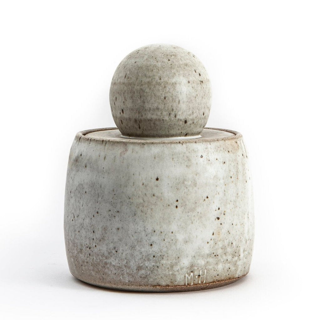 These stoneware jars have a simple, rounded form with a smooth surface. The body of the jar is wide and tapers slightly toward the top, and the lid is crowned with a prominent, round knob that fits comfortably in the hand. The texture of the jar is matte, with a soft, almost velvety feel, and small speckles are present across its surface. The jars feel sturdy and solid, with subtle variations in shape that reflect their handcrafted nature. The lids fit snugly, with the chunky knob providing a tactile featur