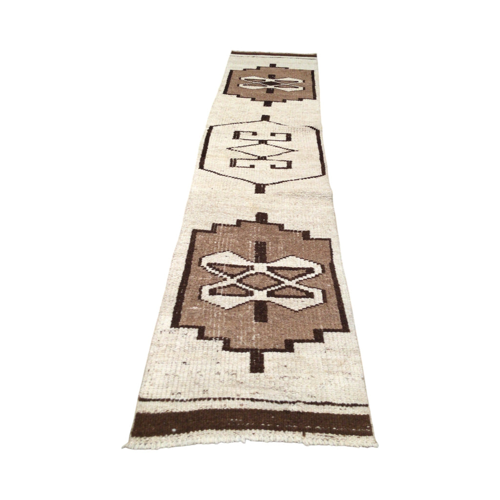 This is a long, narrow rug with a soft, woolen texture. The rug is divided into several sections, each featuring raised, symmetrical patterns with shapes like diamonds and crosses. The colors are muted and natural, blending soft brown and beige, giving it a warm, rustic feel.