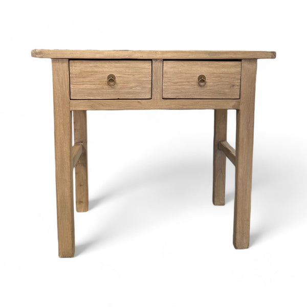 This table is rectangular, made from sturdy, rough-textured wood with a natural finish. It has four thick legs, standing firmly on the ground, and is about waist-high. The front has two drawers, each with a small brass ring handle you can pull, and the overall feel is rustic, as if the wood has been well-worn but carefully crafted. The edges are smooth but the surface has a slight grain that feels natural and earthy.