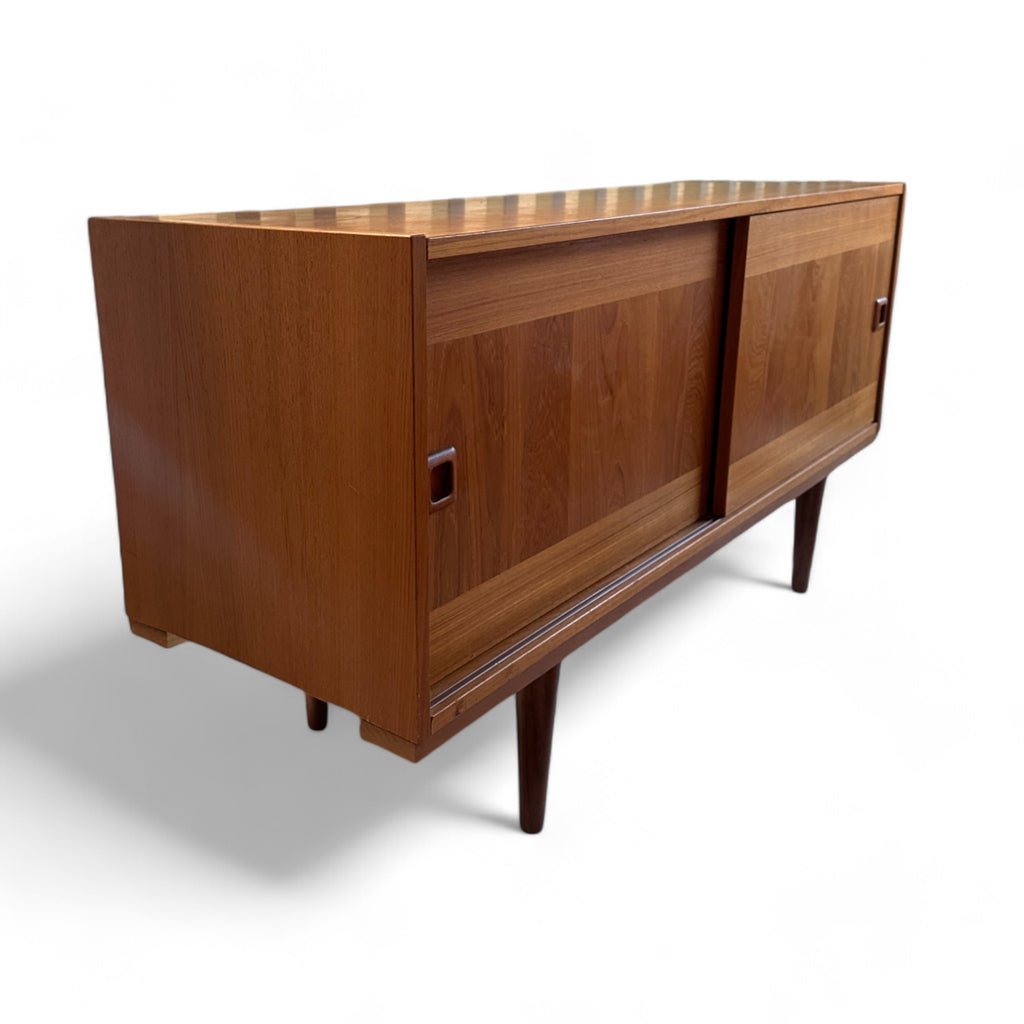 This credenza is a low, rectangular wooden piece with smooth surfaces and clean lines, sitting on four slender, tapered legs that lift it slightly off the ground. The front has two large sliding doors with recessed square handles. The left side behind the door has two shelves, one above the other, while the right side has three tall, narrow columns, ideal for storing things like vinyl records.