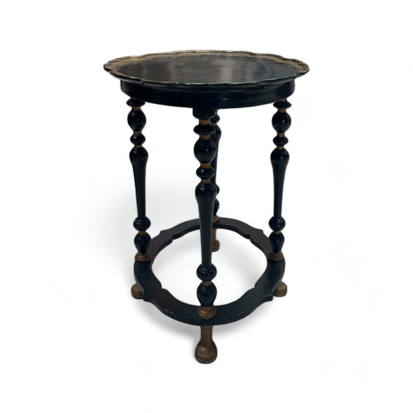 This is a small, oval-shaped side table made from dark ebony wood. The tabletop has a gently scalloped edge with a subtle, decorative gold trim around it. The table stands on four turned legs that are evenly spaced around the edge, connected at the base by a circular support. The legs are slender with a series of rounded, bulging sections that give them a classic, ornate appearance. The table has a delicate and elegant feel, with smooth surfaces and intricate detailing on the legs.