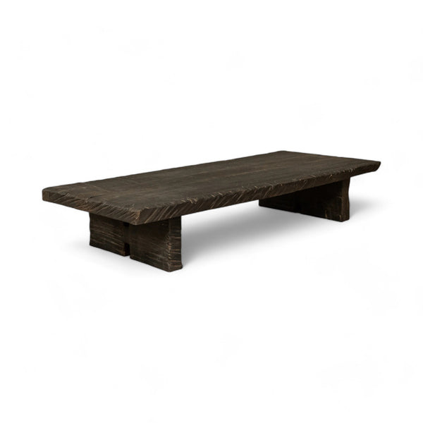 This rectangular coffee table is made of dark, textured wood. The tabletop is thick and flat, supported by two solid, block-like legs on either side. The wood appears rough and natural that add to its rustic, handcrafted appearance.