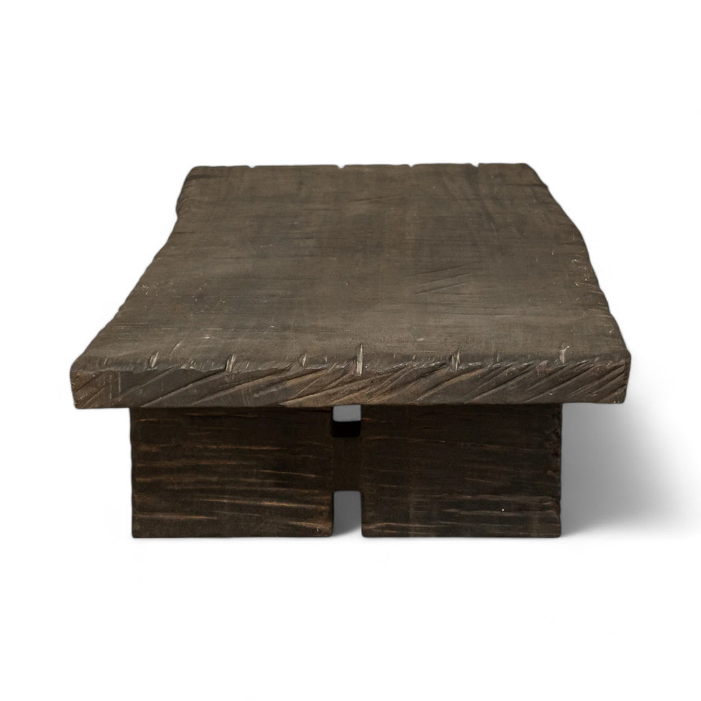 This rectangular coffee table is made of dark, textured wood. The tabletop is thick and flat, supported by two solid, block-like legs on either side. The wood appears rough and natural that add to its rustic, handcrafted appearance.