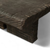 This rectangular coffee table is made of dark, textured wood. The tabletop is thick and flat, supported by two solid, block-like legs on either side. The wood appears rough and natural that add to its rustic, handcrafted appearance.