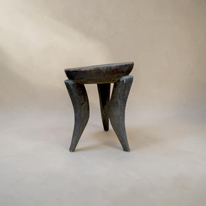 deKor African stool featuring a dark, hand-carved wooden seat and three distinct curved legs on a neutral background.