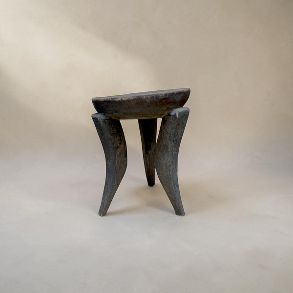 deKor African stool featuring a dark, hand-carved wooden seat and three distinct curved legs on a neutral background.