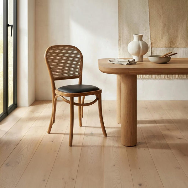 deKor rattan dining chair with a natural wood frame, woven cane backrest, and a dark polyurethane seat cushion.