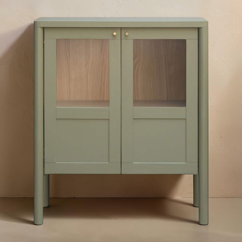 deKor sage green cabinet with tempered glass doors, solid pine frame, and minimalist bronze hardware.