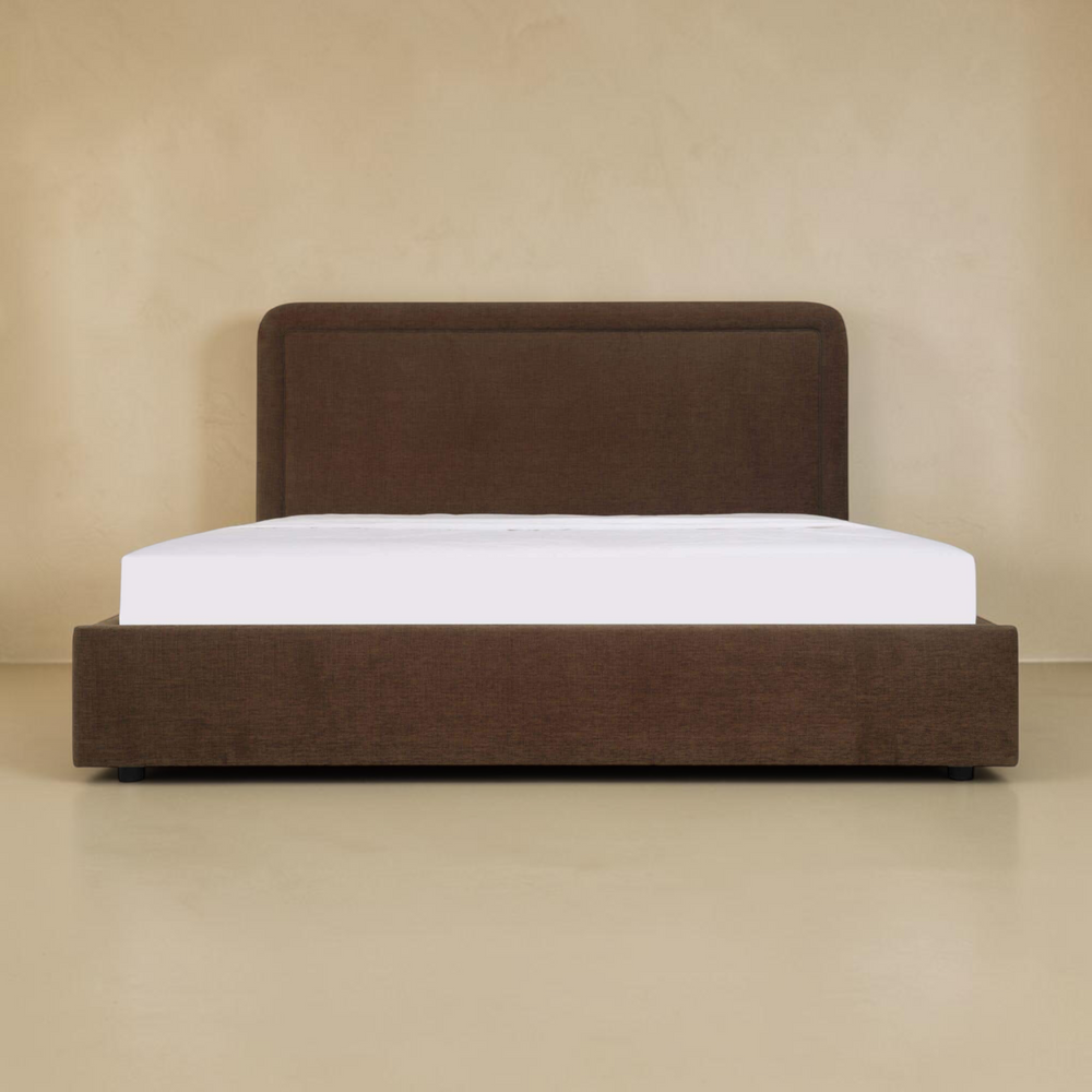 deKor Simon storage bed in dark brown fabric with a rounded headboard and piped detailing, front view.