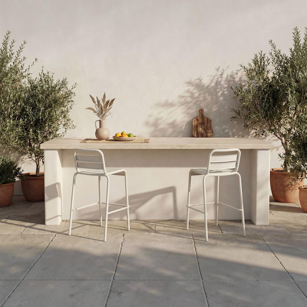 Two deKor white slatted steel outdoor bar stools positioned at a modern stone bar table on a sunlit patio.