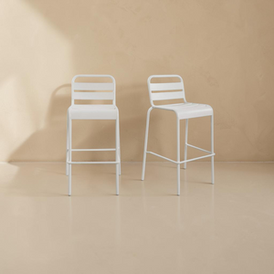 deKor set of two white slatted steel outdoor bar stools with a modern, minimalist design against a neutral wall.