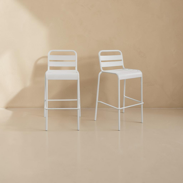 deKor set of two white slatted steel outdoor bar stools with a modern, minimalist design against a neutral wall.