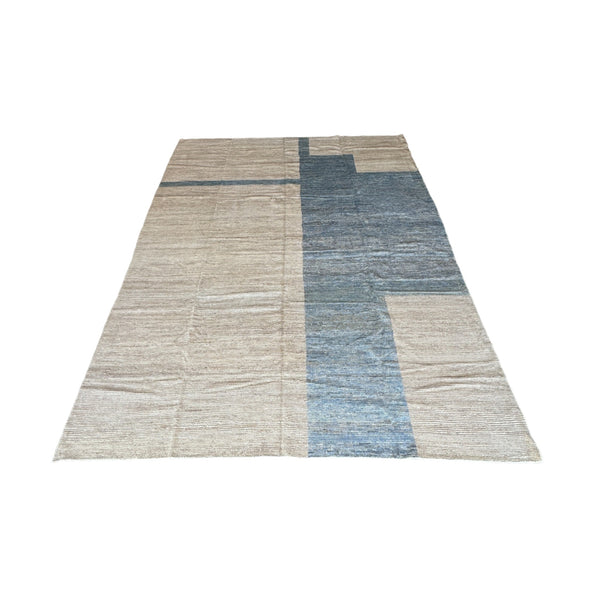 This rug has a smooth, soft texture, crafted from wool. The design is minimalistic with large, simple geometric shapes. The main color is a warm, neutral beige, with some areas of the rug featuring a cool blue. The blue sections form an abstract, blocky pattern, which contrasts subtly with the beige background.