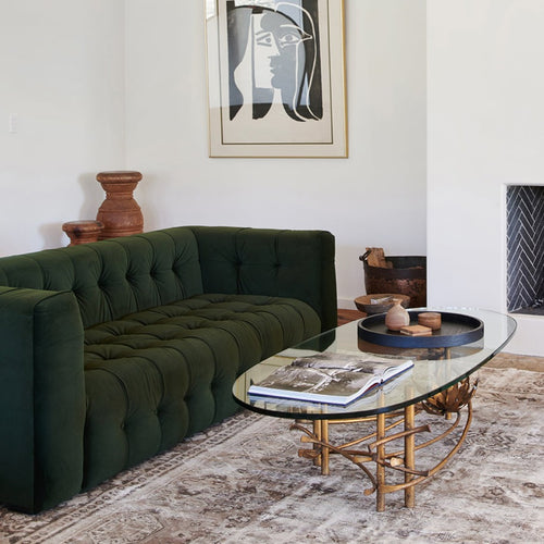 Green tufted velvet sofa in white room with vintage glass and guilded coffee table and abstract art on wall. 