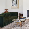 Green tufted velvet sofa in white room with vintage glass and guilded coffee table and abstract art on wall. 